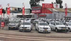 מגרש רכבים פתוח מלא ברכבים מסוגים שונים, חונים באופן מסודר תחת שמיים בהירים.